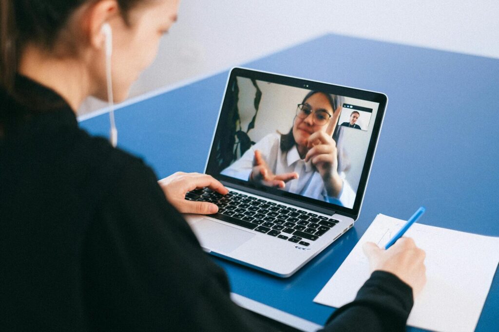 A person is seen speaking to another virtually through a laptop on a blue tabletop while taking notes, showing how a licensed therapist would operate through a telehealth platform while delivering outpatient therapy services.