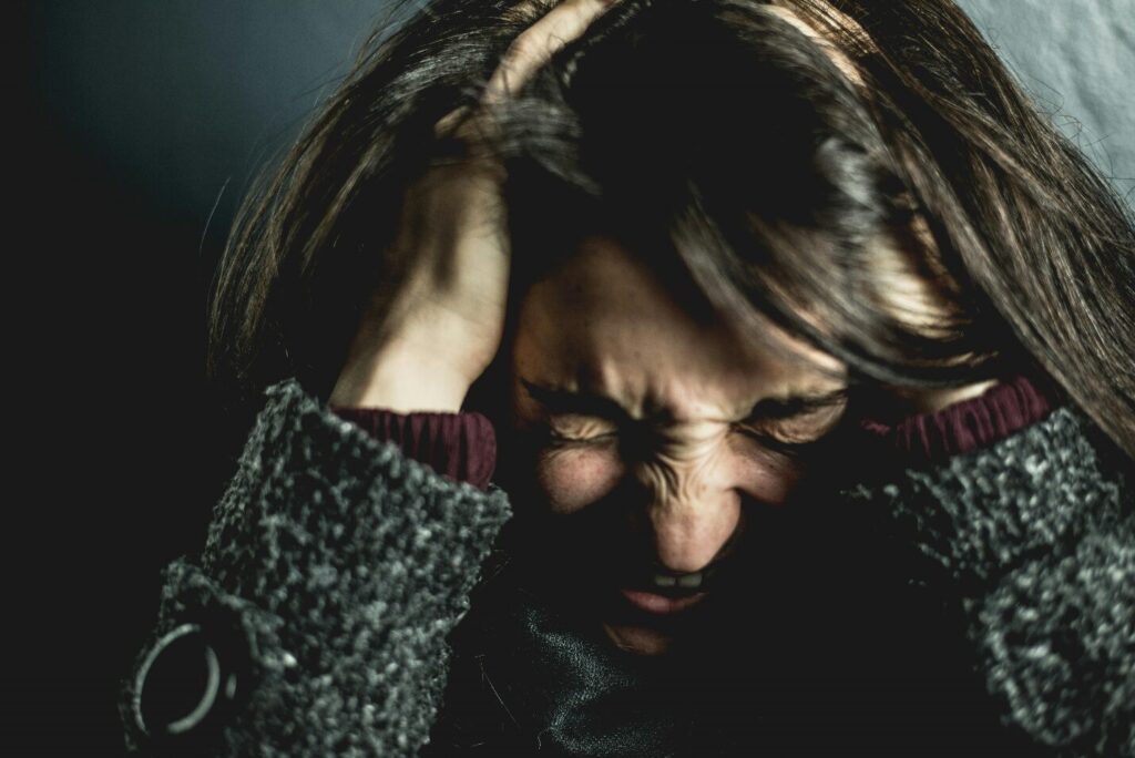A woman holds her head while squeezing her eyes tight and her mouth slightly open while looking down in a dark image, showing the importance of anxiety management techniques to regulate emotions and maintain composure.