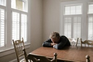 A man slumps onto a table with his face resting on his arms crossed on the tabletop surrounded by chairs and windows with shutters, highlighting the signs that mental health therapy may be beneficial to treat anxiety or depression.